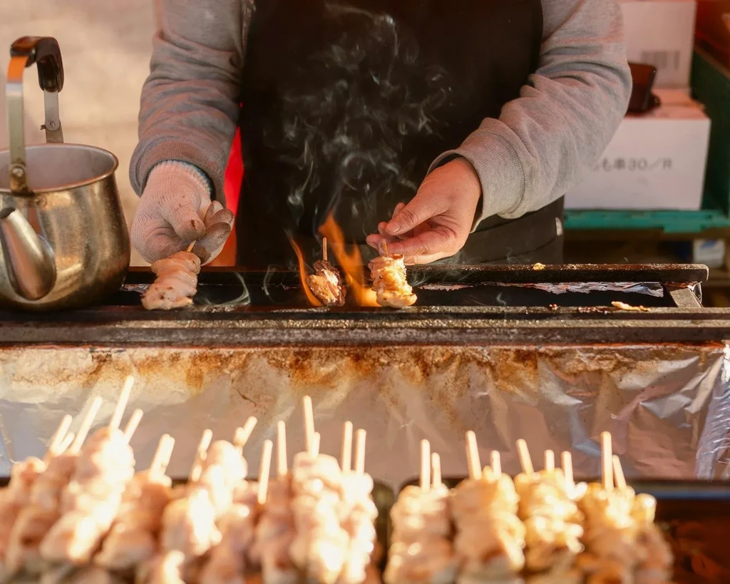 Gloved hands turn yakitori skewers on a flaming grill.