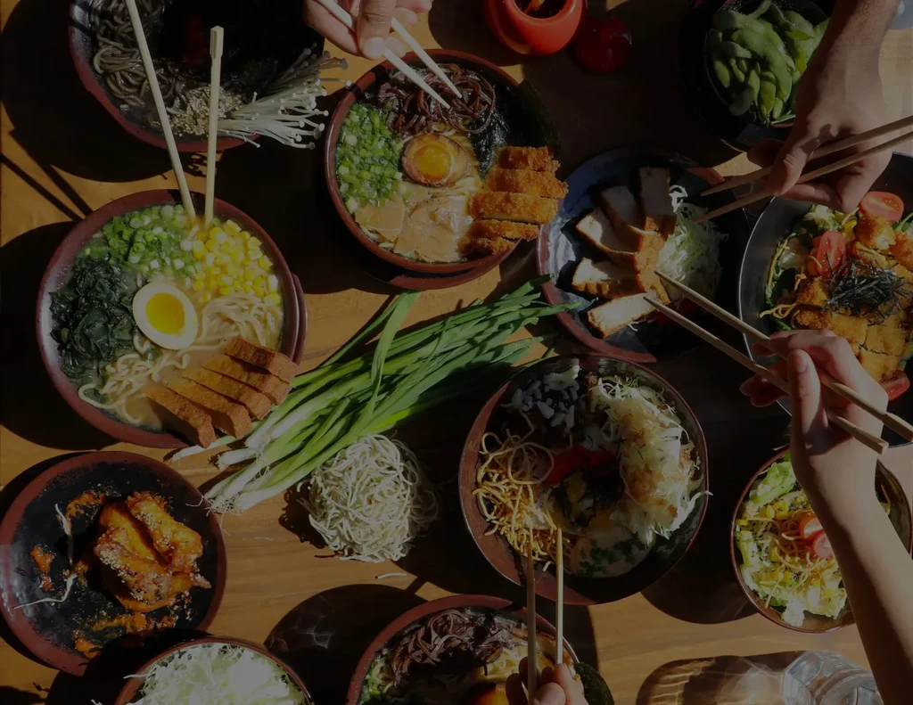 Hands sharing vibrant ramen and Asian dishes on a wooden table.