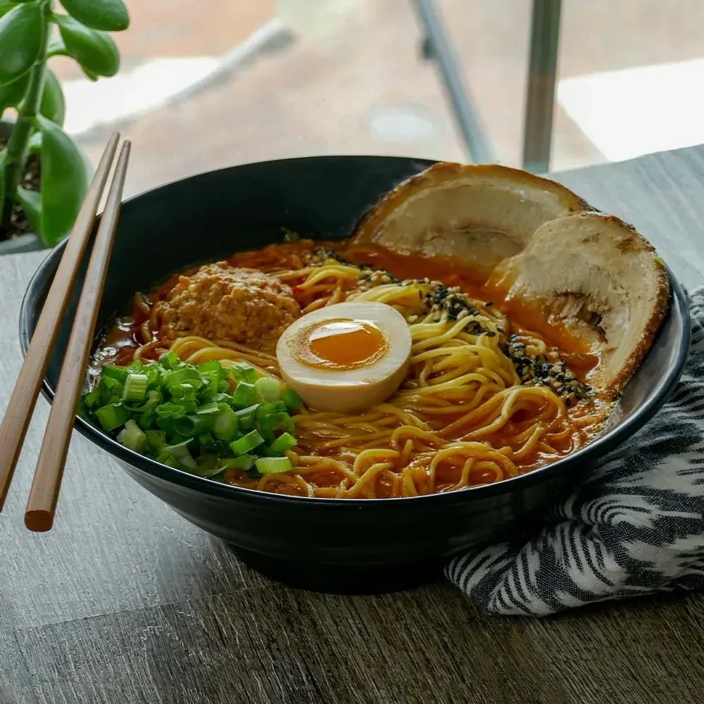 Close-up of ramen bowl with noodles, egg, pork, green onions, chopsticks.