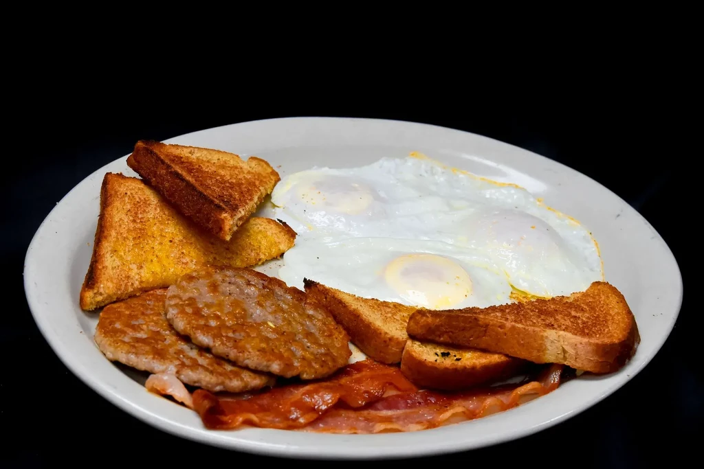 Breakfast plate featuring fried eggs, crispy bacon, sausage, and golden toast.