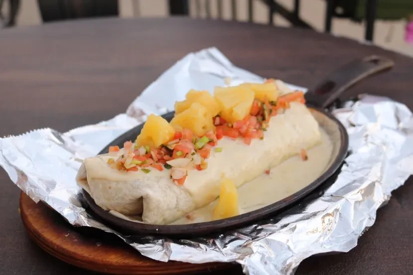 Burrito in skillet, covered with creamy sauce, tomato salsa, and pineapple.