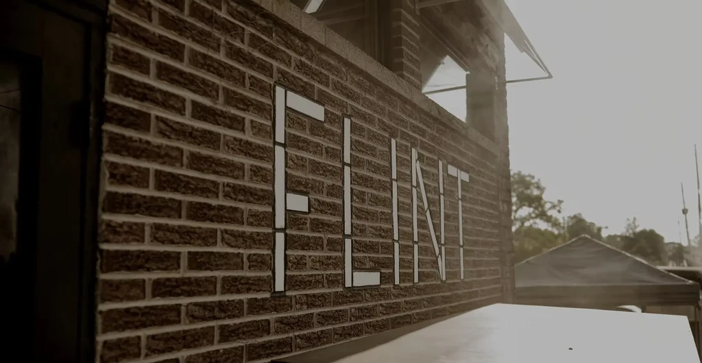 White block letters spell "FLINT" on a dark brown brick wall.