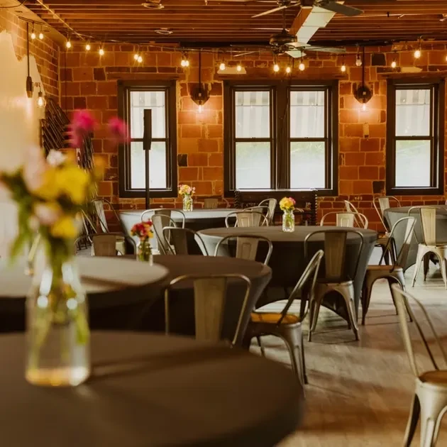 Rustic event space with round tables, brick walls, wooden beams, string lights.