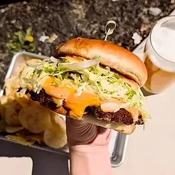 Hand holding a large burger, chips, and a foamy beverage.