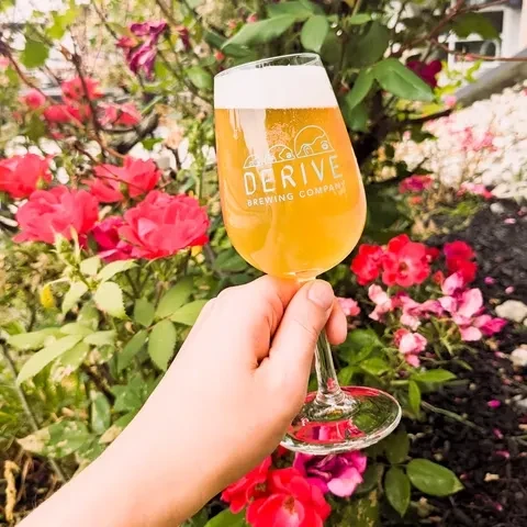 Person's hand holds a "DERIVE" beer in a flowery garden.