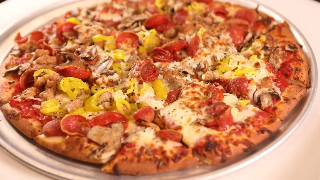 Round pizza with pepperoni, sausage, mushrooms, yellow peppers on a metal pan.
