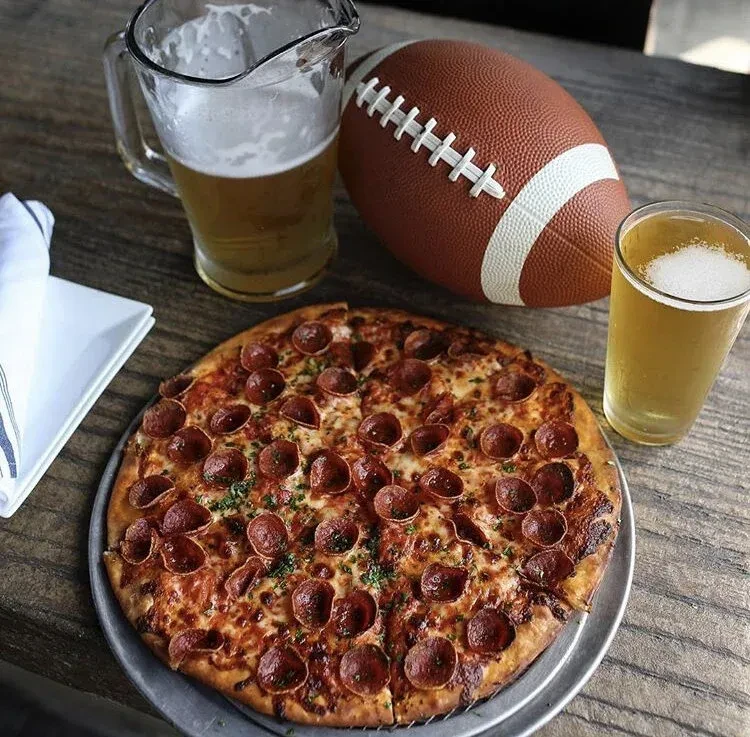 Top-down of pepperoni pizza, beer, football on rustic wooden table.