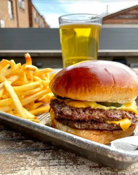 Double cheeseburger, fries, and beer on rustic tray, outdoor setting.