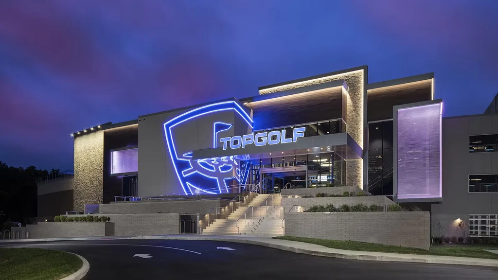 Exterior of a modern Topgolf facility glowing under a dramatic twilight sky.