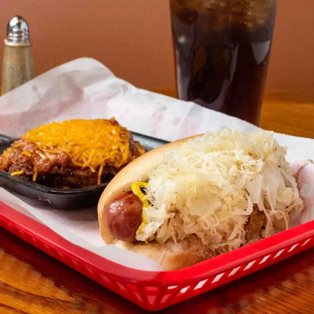 Hot dog with sauerkraut, mustard, and chili cheese in a red basket.