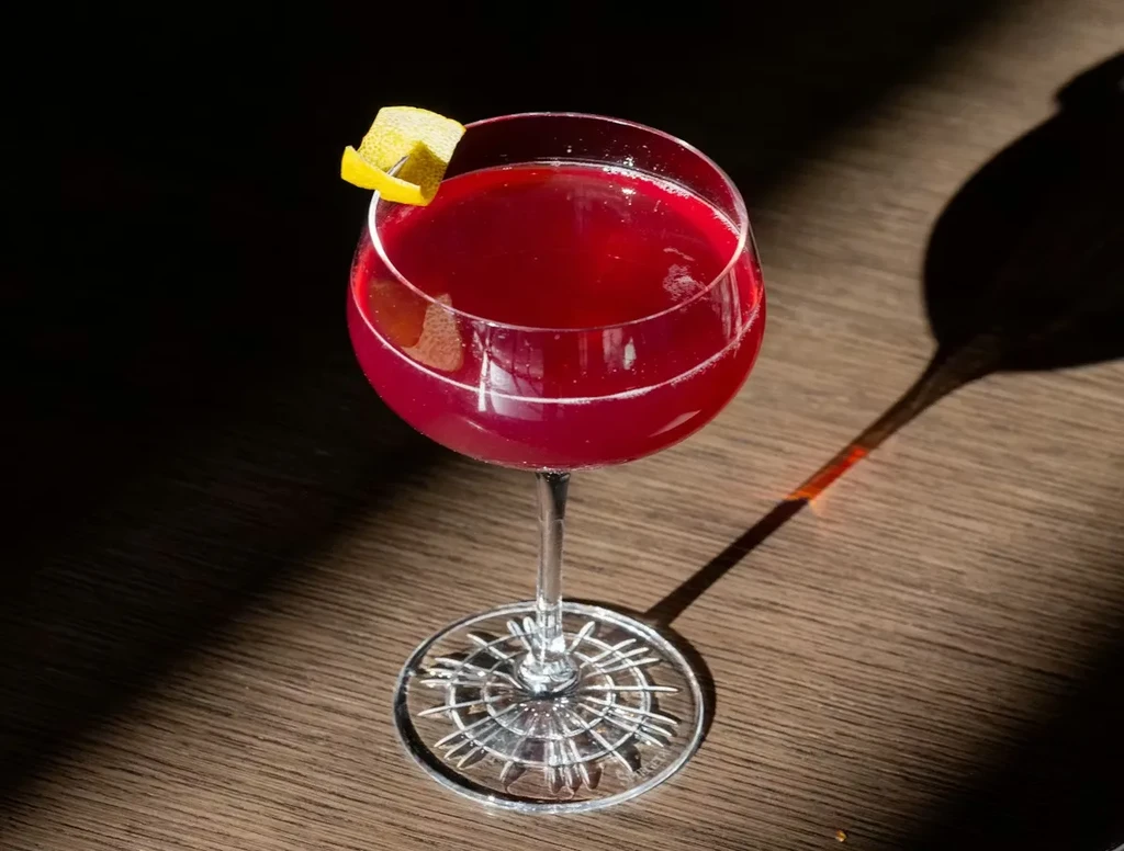 Deep red cocktail in stemmed glass with lemon peel and long shadow.
