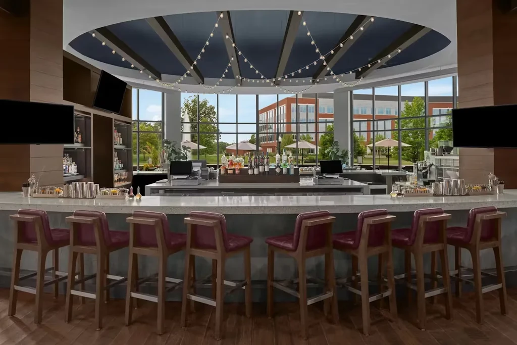 Bright modern bar, maroon stools, stocked liquor, large windows, outdoor view.