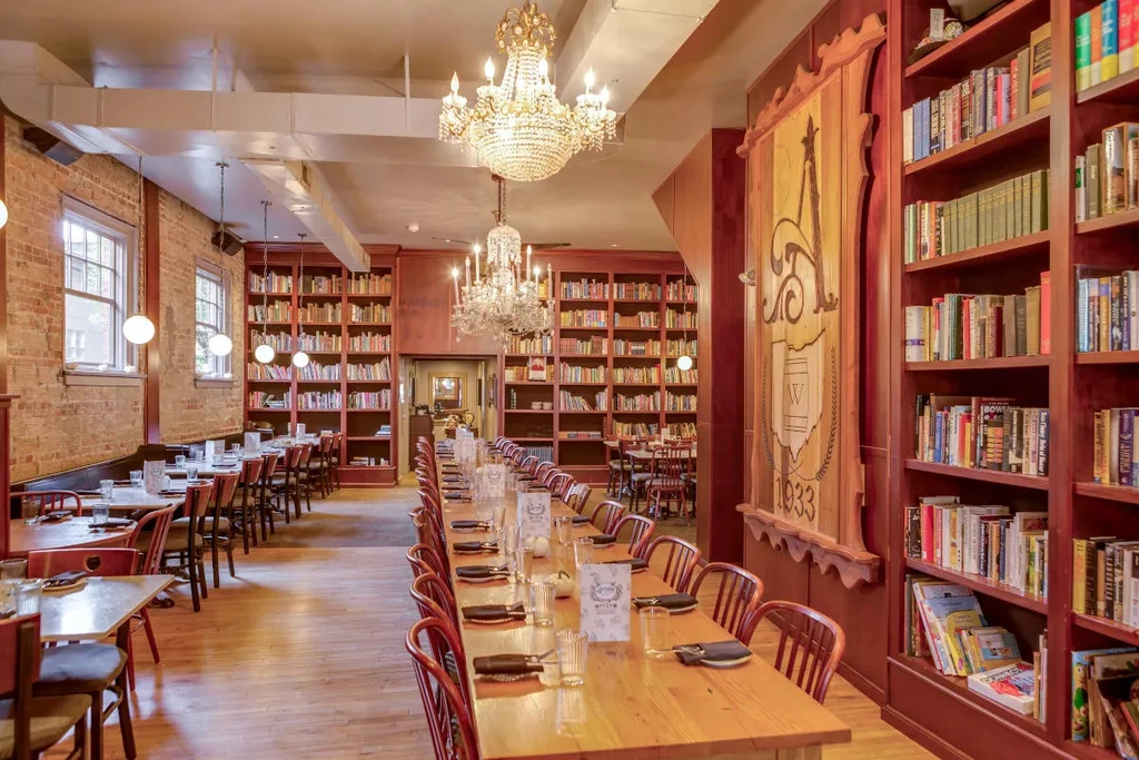 Library-themed restaurant interior with bookshelves, brick wall, and communal dining table.