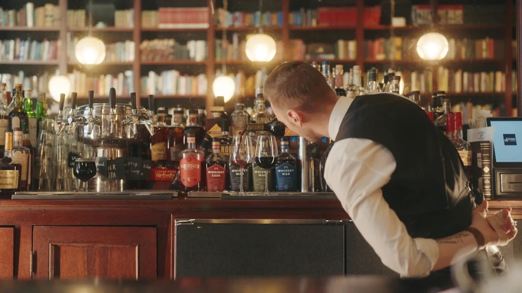 Bartender pours drink at a wooden bar counter with bookshelves.