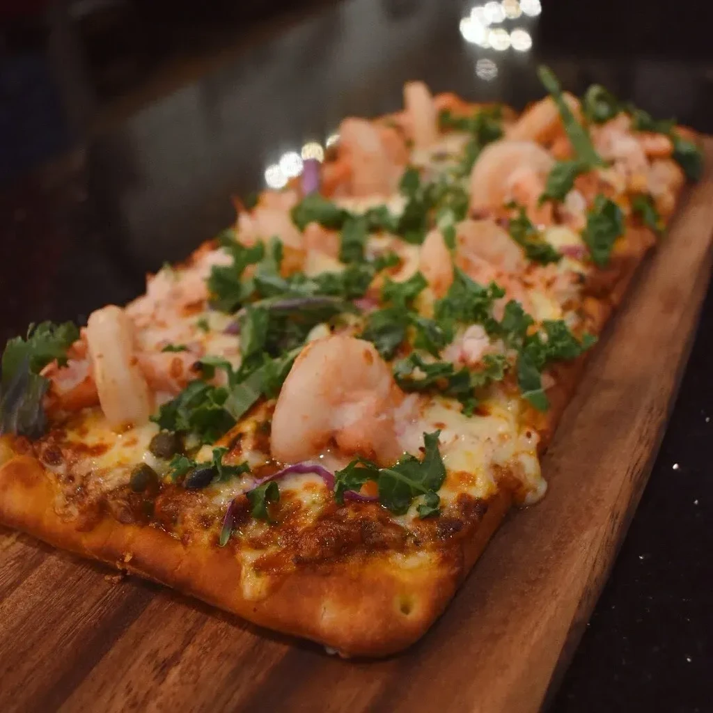 Shrimp, kale, melted cheese flatbread pizza on a rustic wooden board.