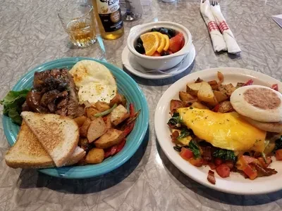 Tabletop breakfast spread with grilled meat, eggs, potatoes, fruit, and drinks.