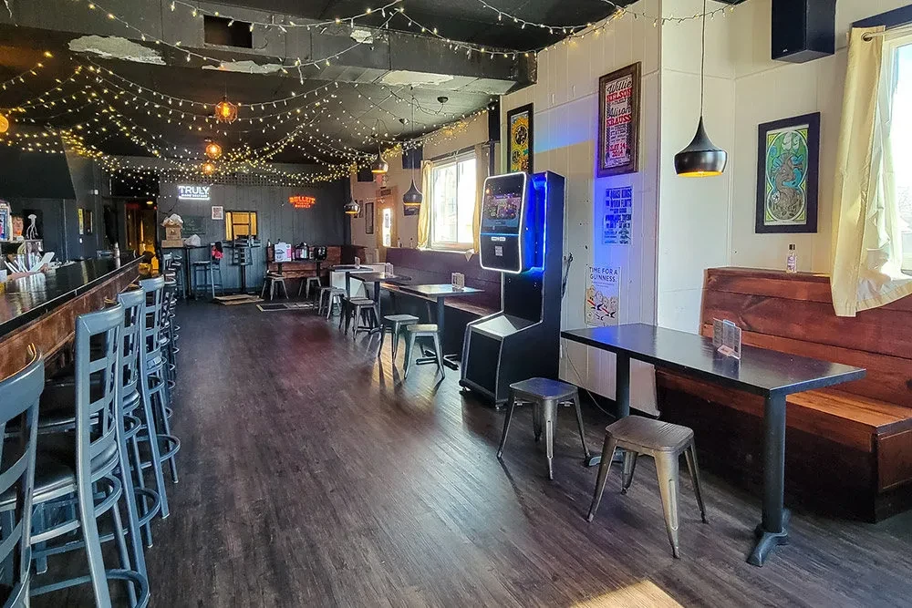 Cozy bar interior with wooden bar, booths, string lights, and arcade machine.