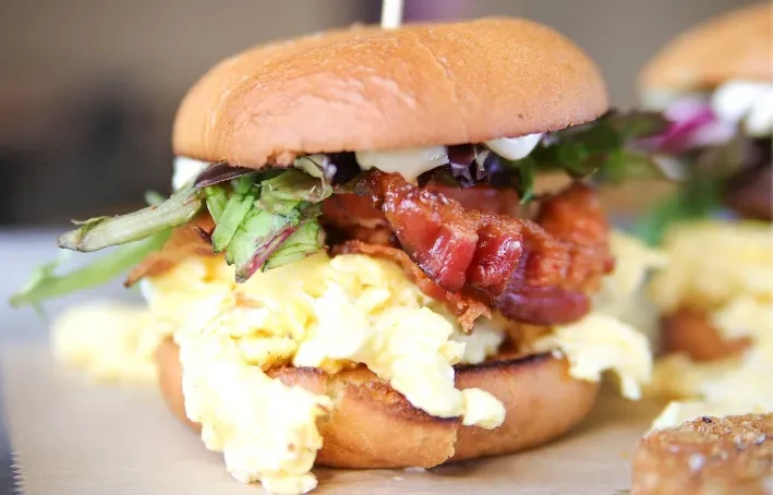 Golden bun breakfast sandwich with eggs, bacon, greens, and sauce.