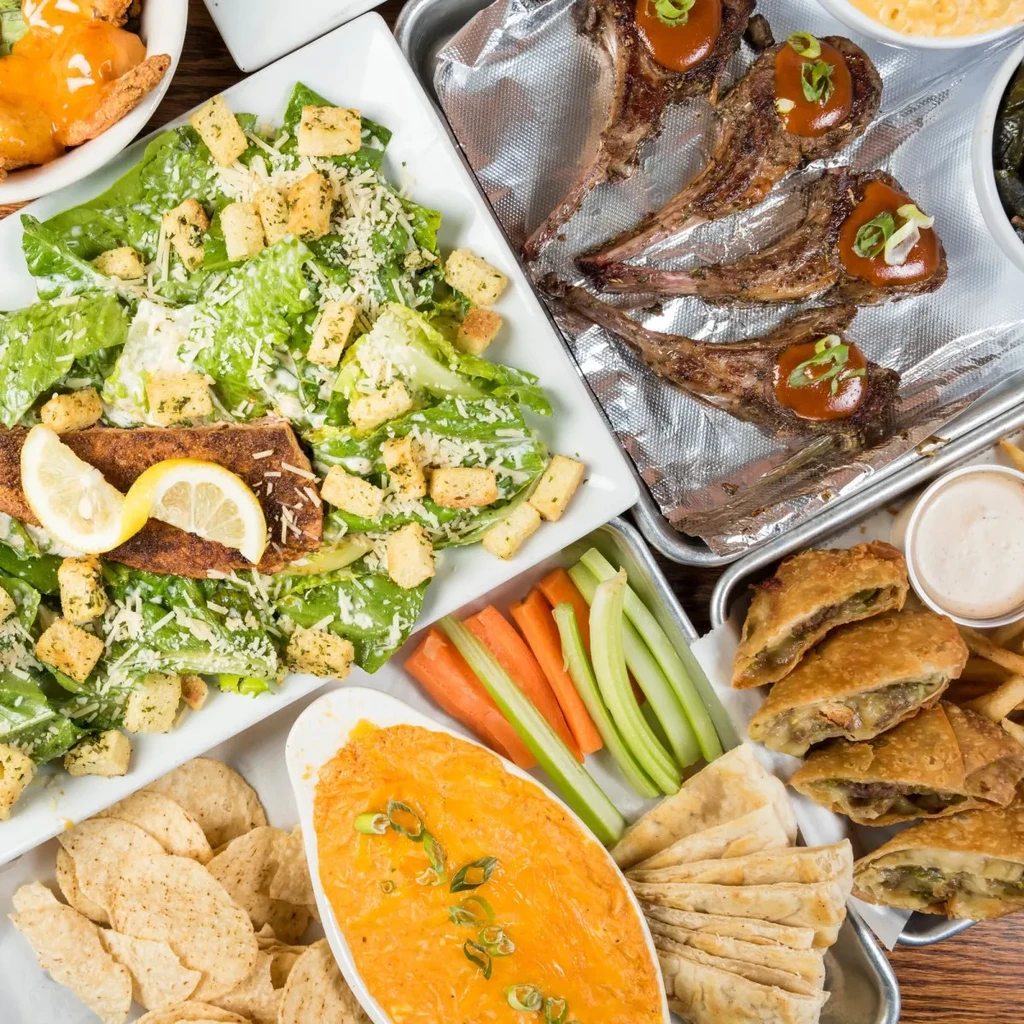Overhead view of diverse dishes: salmon salad, lamb chops, fried chicken, dips.