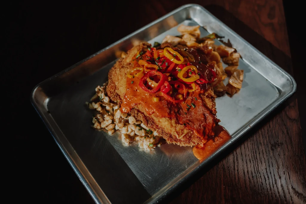 Plated breaded item with orange sauce, chili, herbs, and grains.