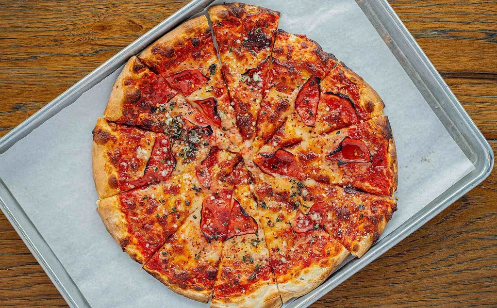 Golden pizza, sliced, with cheese, red toppings, and herbs on metal tray.