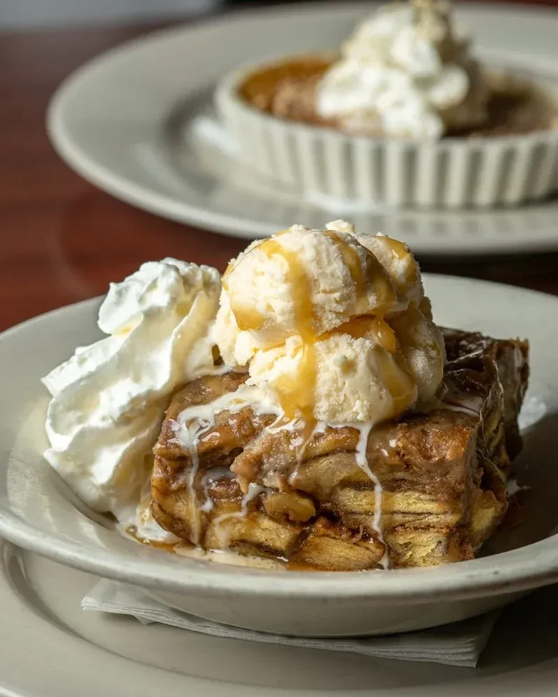 Bread pudding with ice cream, caramel, whipped cream; blurred custard in background.