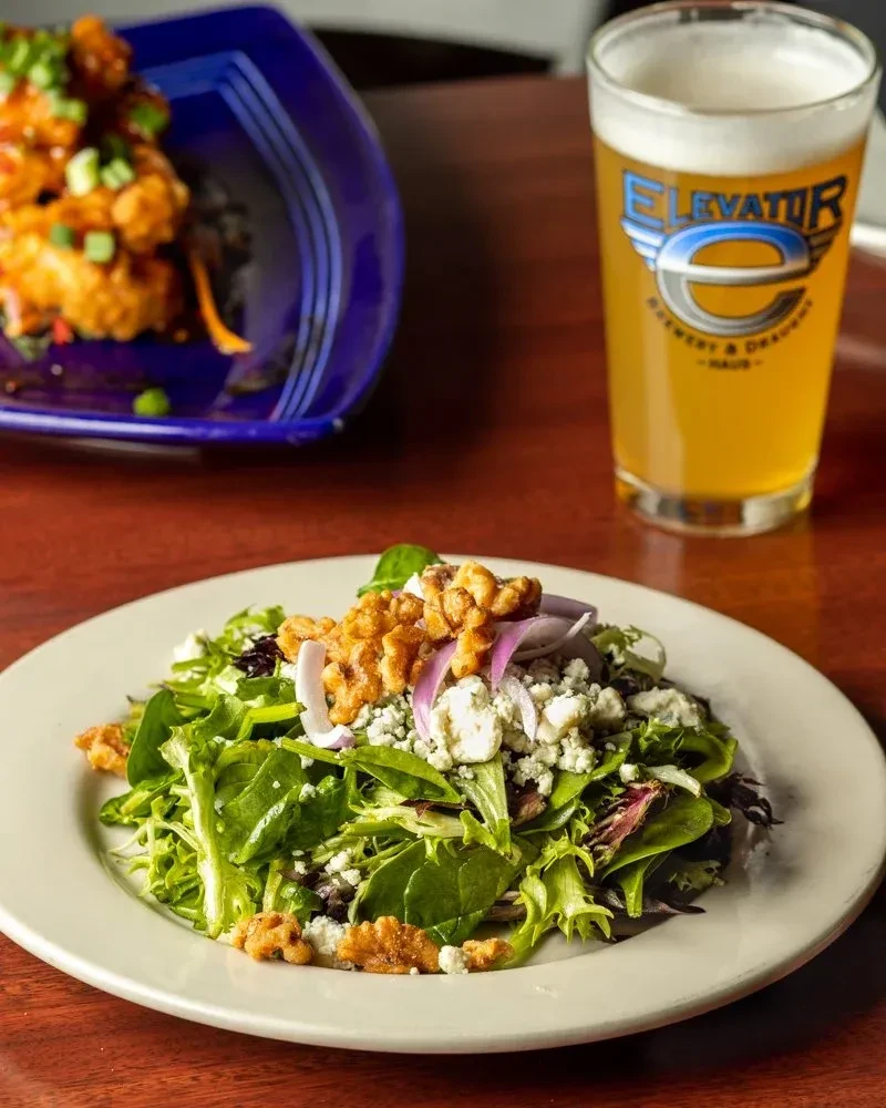 Salad, sauced bites, and Elevator Brewery beer on a dark table.