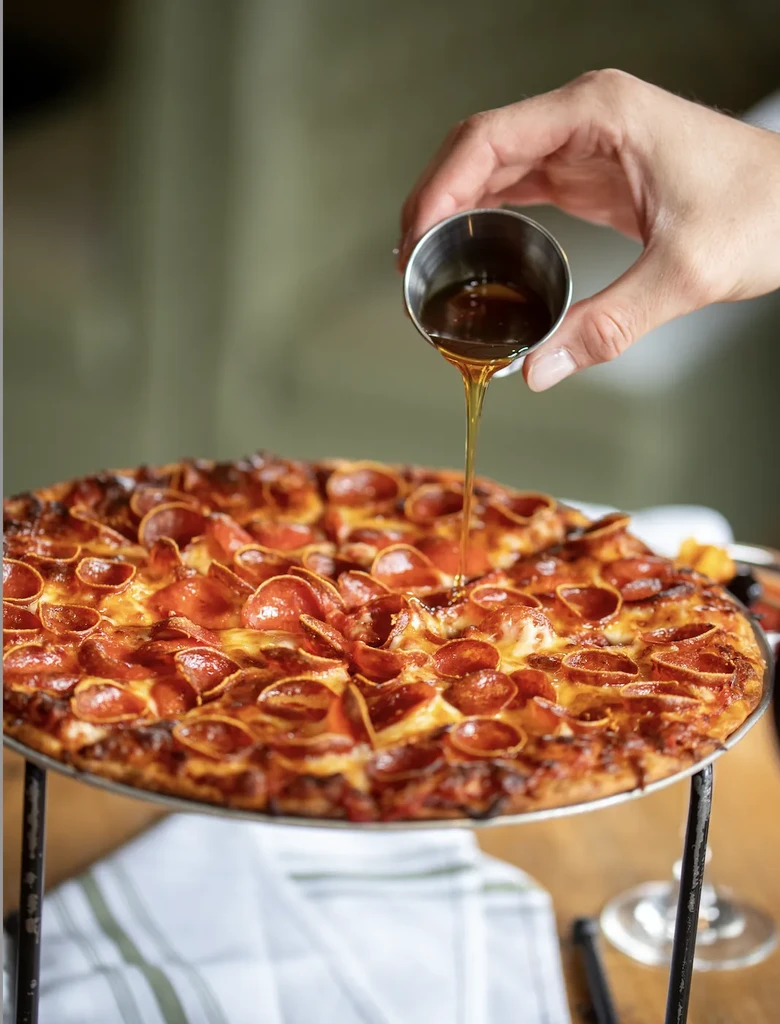 Hand drizzles amber liquid over crispy pepperoni pizza.