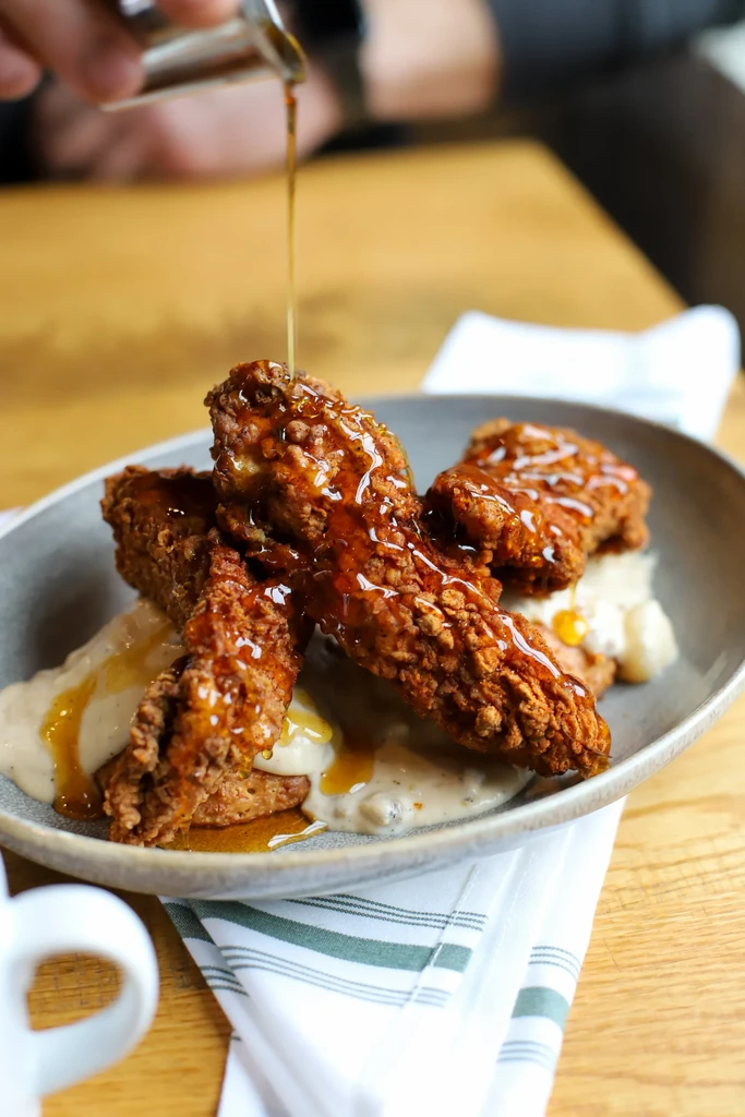 Hand pours amber syrup over fried chicken tenders and creamy gravy.