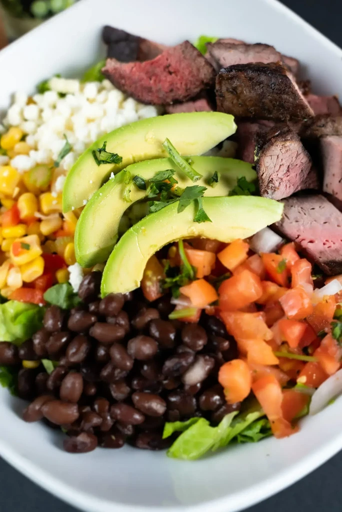 White bowl filled with grilled steak, avocado, corn, beans, and tomatoes.