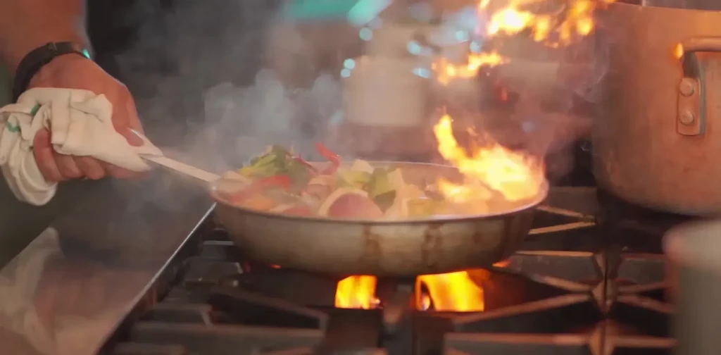 Chef's hands stir-frying vegetables in a pan over gas flames.