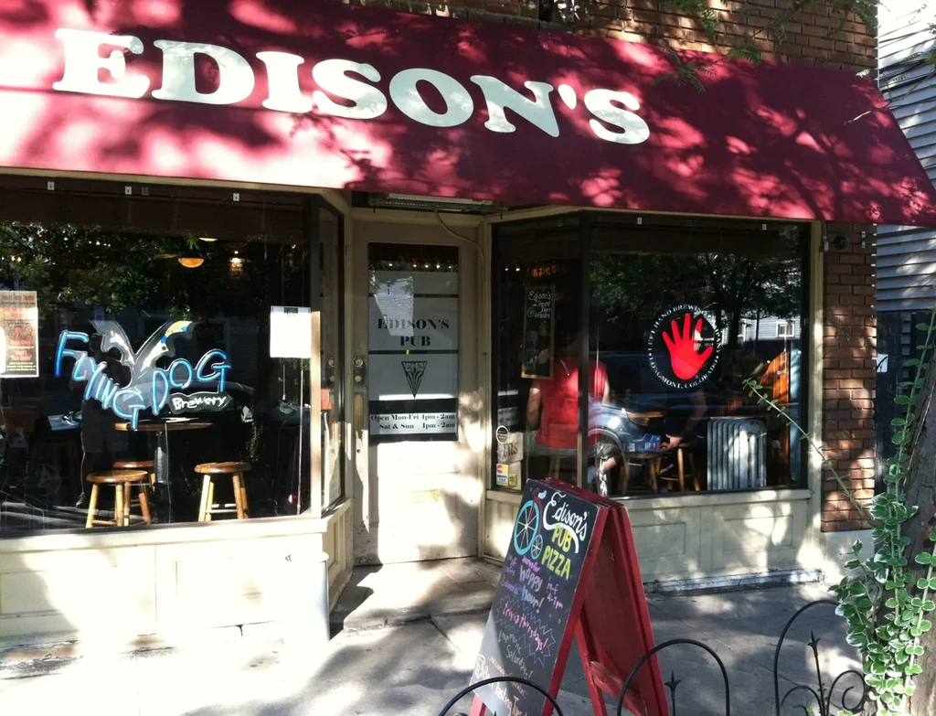 Sunlit exterior of Edison's Pub with brewery signs and pizza chalkboard.