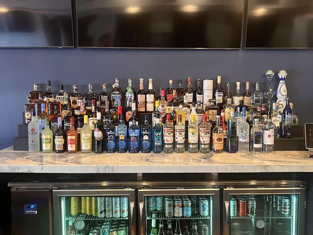 A well-stocked modern bar with liquor bottles, refrigerated drinks, and wall screens.