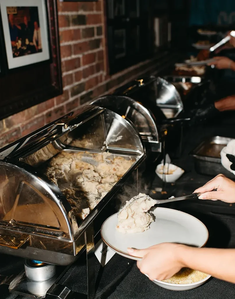 Person's hands serving mashed potatoes from a buffet chafing dish.