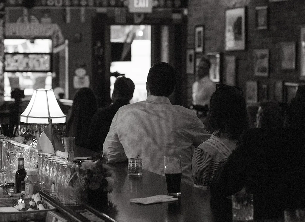 Black and white photo: patrons seated at a dimly lit bar counter.
