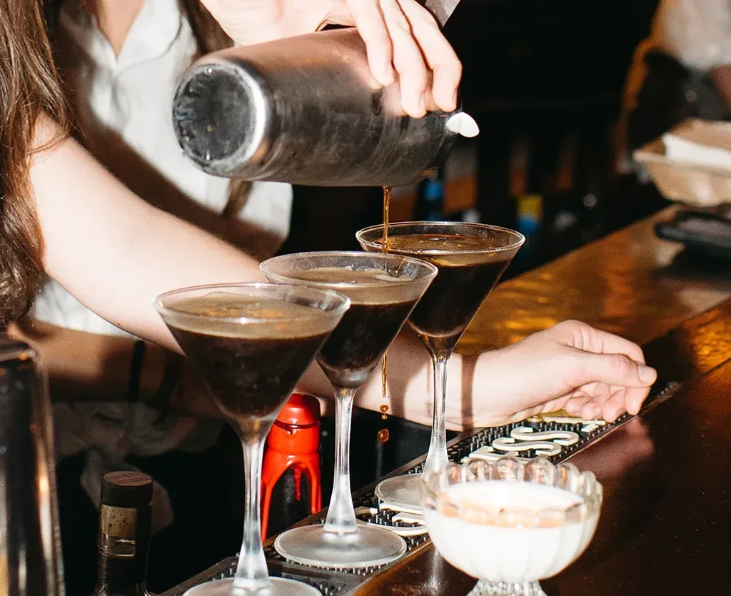 Bartender pours dark cocktail from shaker into three martini glasses.