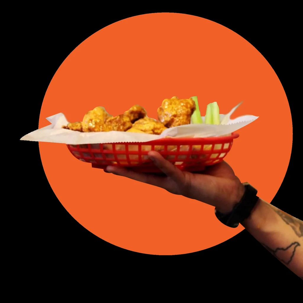 Hand holding a red basket of saucy chicken wings and celery.