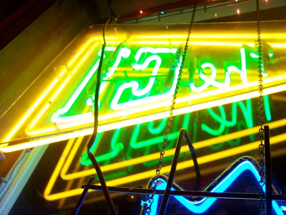 Green cursive neon sign, yellow geometric frame, and blue pattern.
