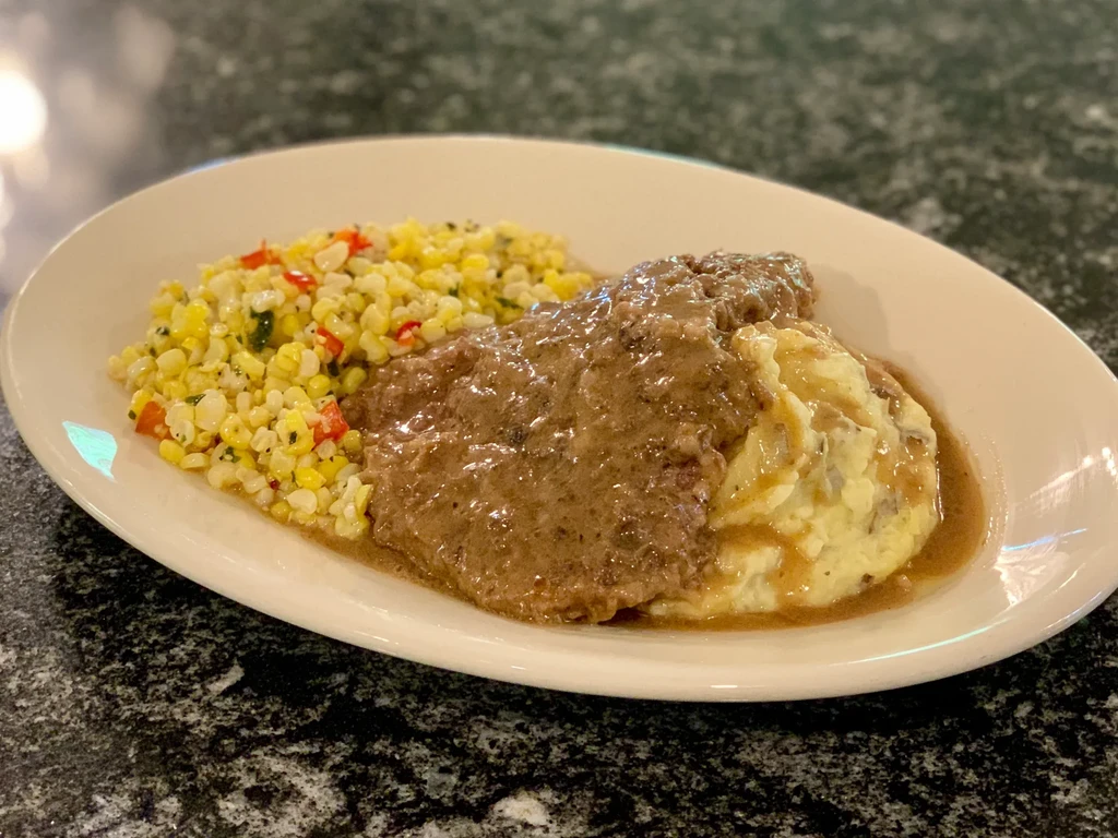 Smothered steak, corn salad, and mashed potatoes on an oval plate.