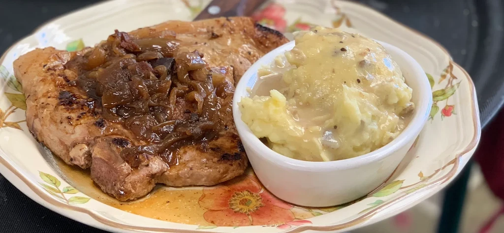 Grilled pork chop, caramelized onions, mashed potatoes on floral plate.
