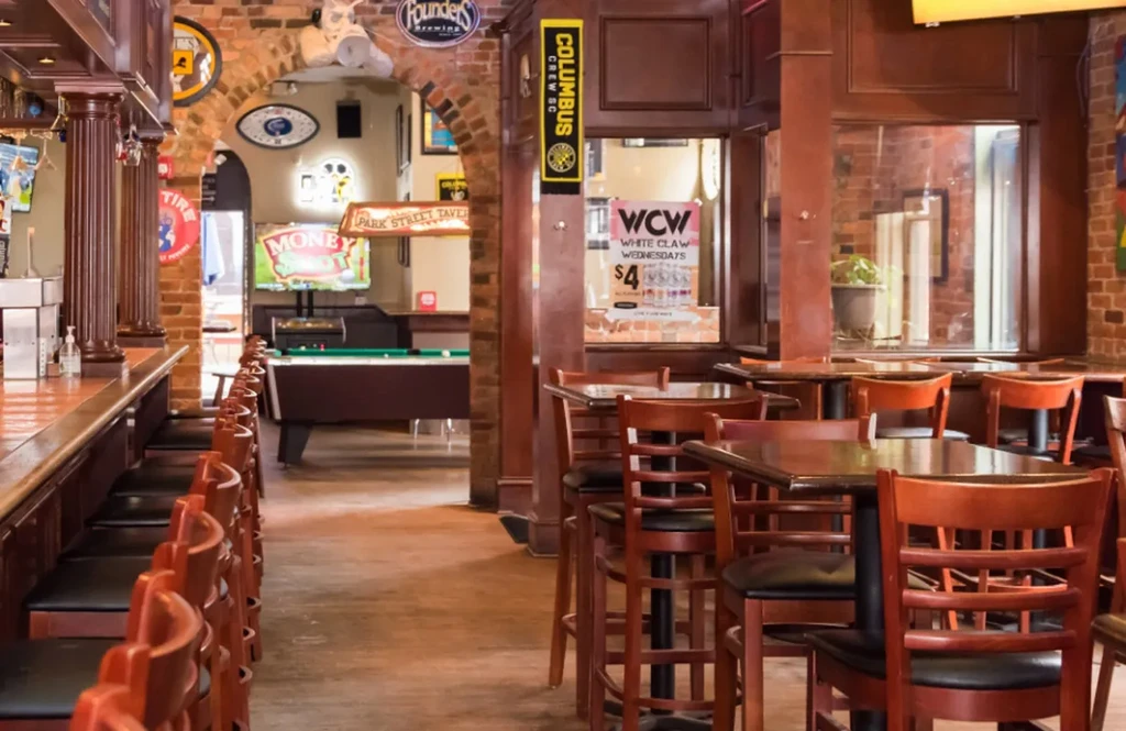 Rustic bar interior: wooden bar, high-top tables, and visible pool room.