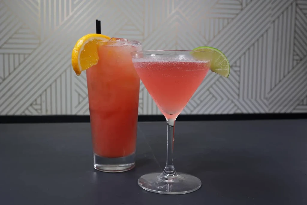 Vibrant red-orange and pink cocktails, garnished, in front of geometric wallpaper.