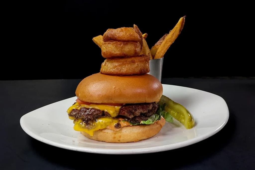 Double cheeseburger with onion rings, potato wedges, and pickle on white plate.