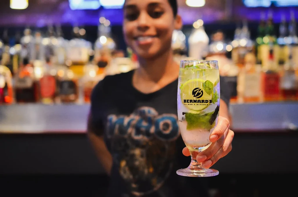 Hand holds a mojito cocktail with ice and Bernard's Bar logo.
