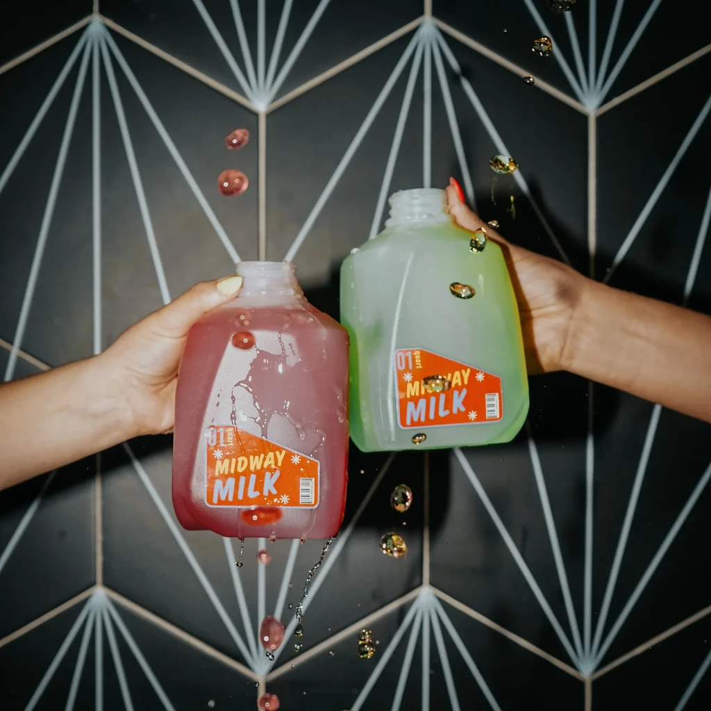 Two hands hold splashing bottles of Midway Milk, pink and green.