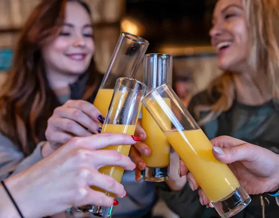 Four manicured hands clinking glasses with yellow-orange drinks.