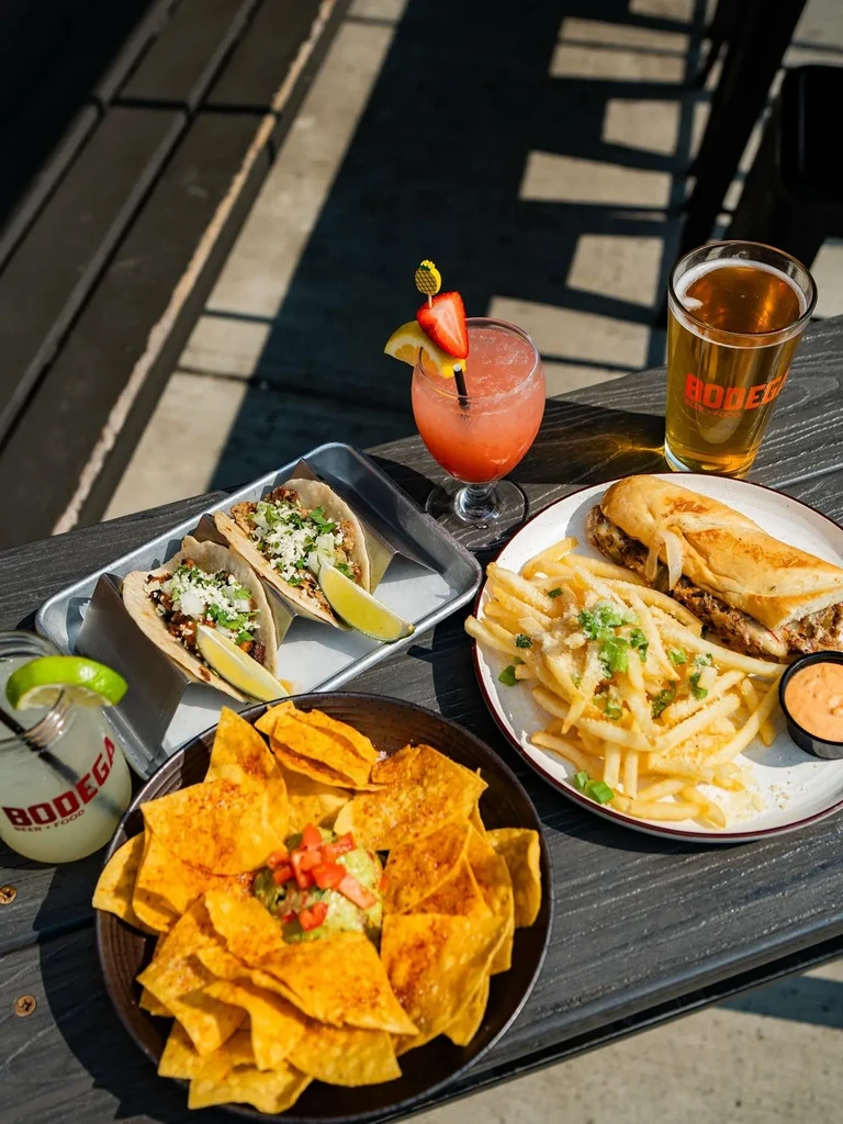 Tacos, guacamole, sandwich, fries, and Bodega drinks on outdoor table.