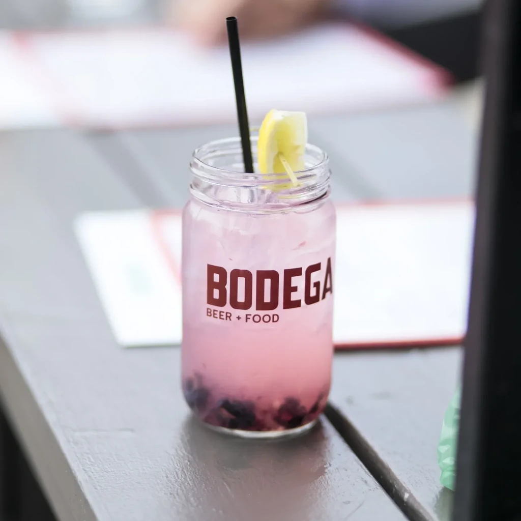 Pink beverage with ice, berries in BODEGA jar, lemon, black straw.