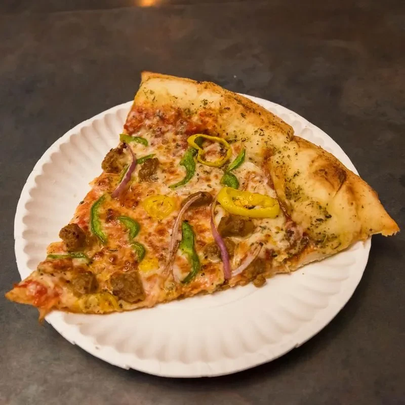 Pizza slice with sausage, peppers, onions, and cheese, on paper plate.