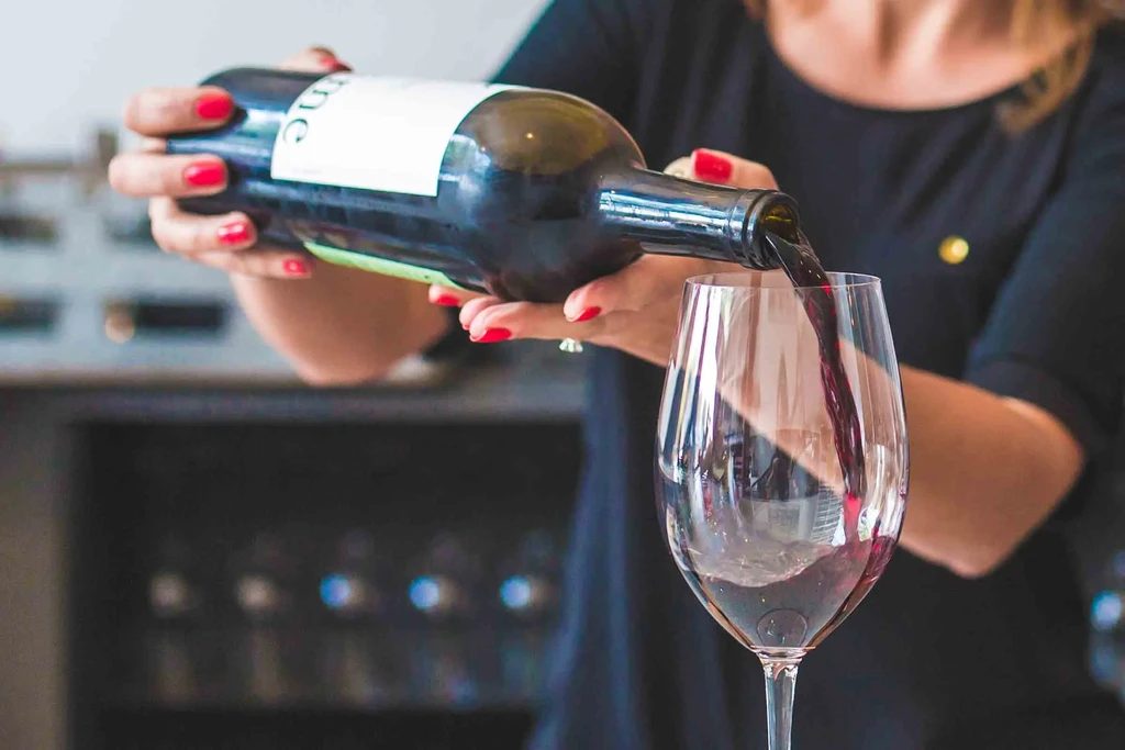 Red-nailed hands pour red wine from a bottle into a glass.